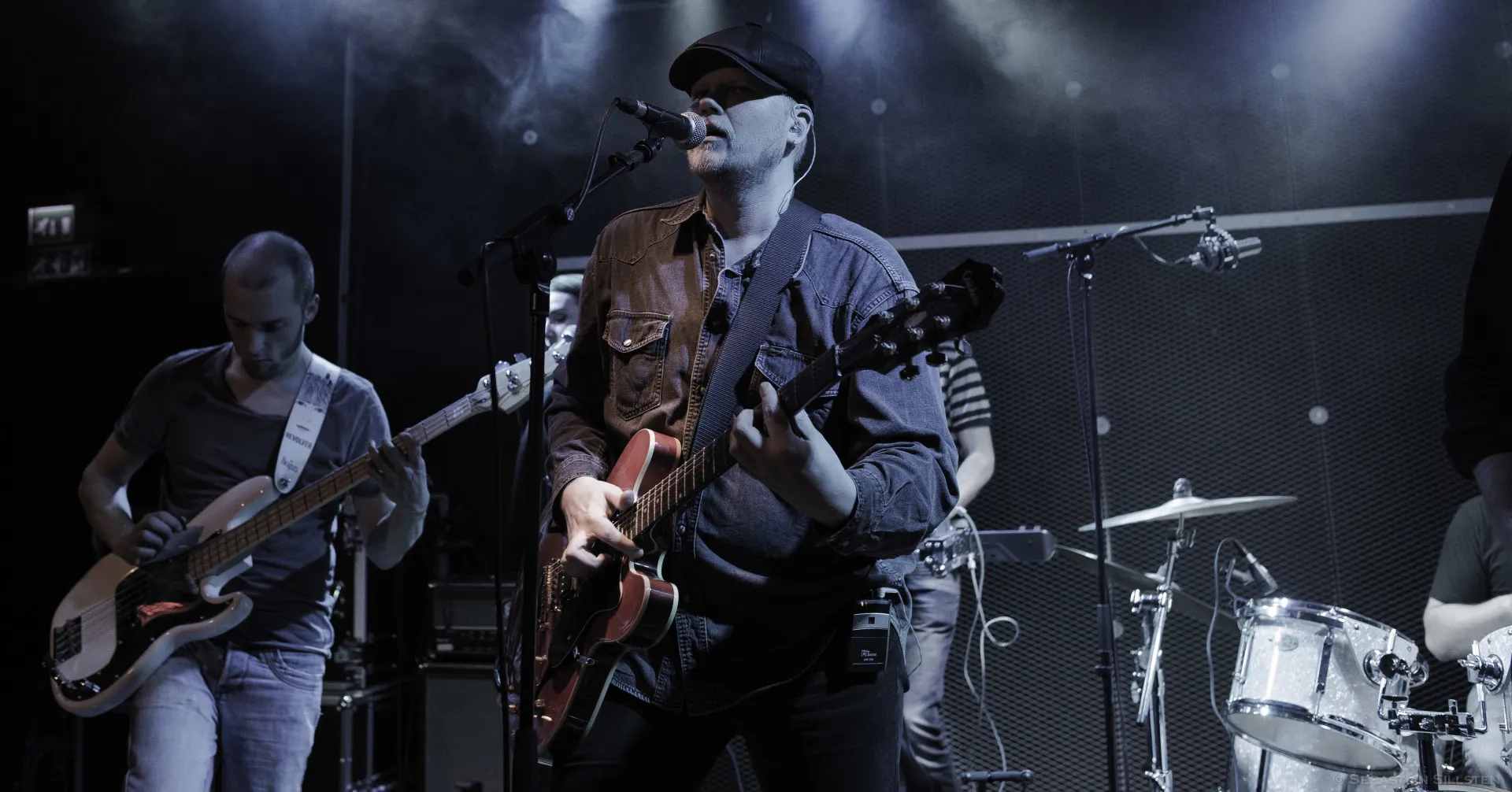 Hewey singing and playing guitar on stage, the bassist behind him